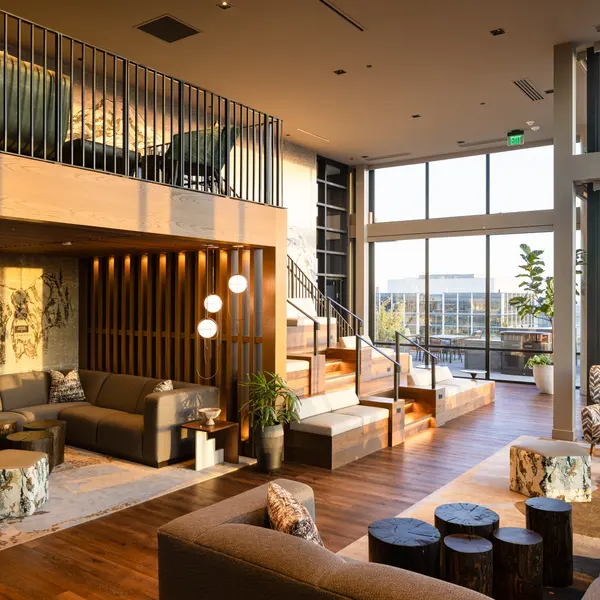 A stylish and modern lounge area featuring a cozy seating arrangement, large windows letting in natural light, and a wooden structure with a staircase leading to an upper level. Decorative elements include plants and wooden accents, creating a warm and inviting atmosphere.
