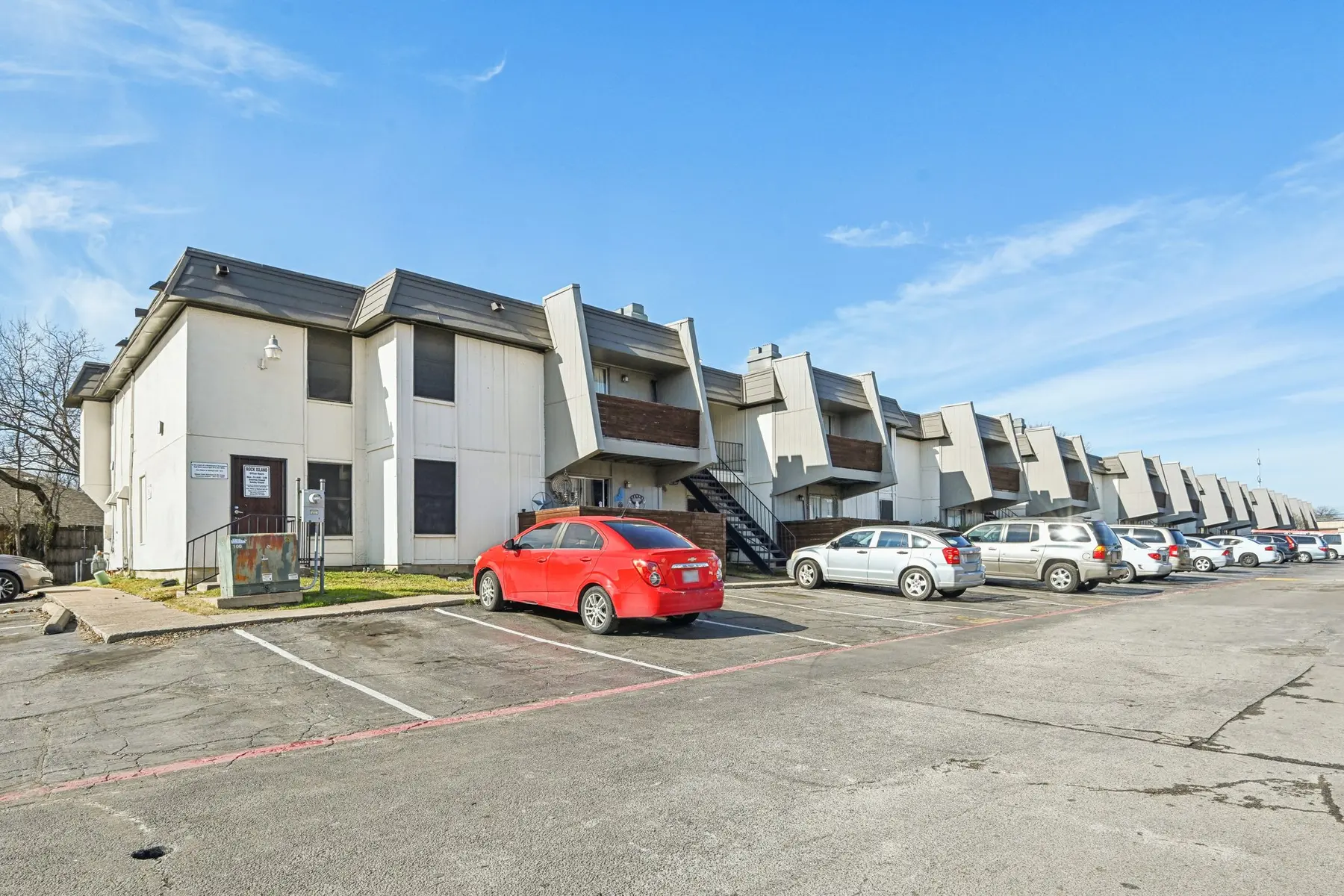 View of a multi-unit apartment complex with a row of parked cars in front and a clear blue sky above.