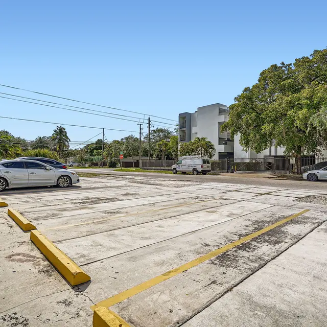 A wide view of a mostly empty parking lot with a few cars parked along the sides. Trees and buildings are visible in the background under a clear blue sky.