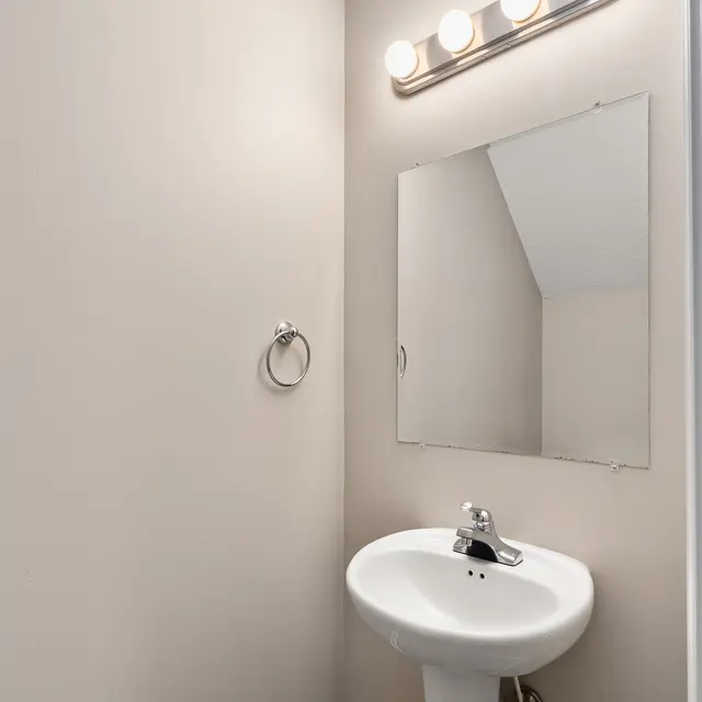 A small bathroom with beige walls, featuring a pedestal sink and a large wall-mounted mirror. Above the mirror, there are four light bulbs on a fixture. A towel ring is mounted on the wall nearby.