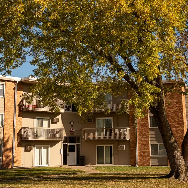 A two-story apartment building surrounded by trees and grass. The building has a brick facade and several balconies. Autumn leaves can be seen on the tree in front of the building.