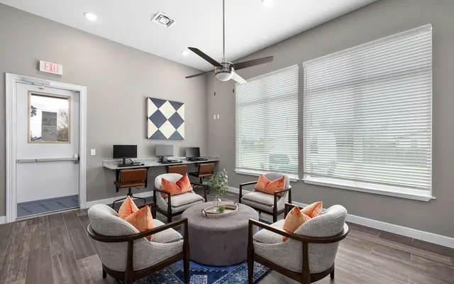 A modern office space featuring four comfortable chairs with orange cushions arranged around a circular table. Two desks with computers are visible against the wall, and large windows with horizontal blinds provide natural light. A geometric wall art piece hangs above the desks.