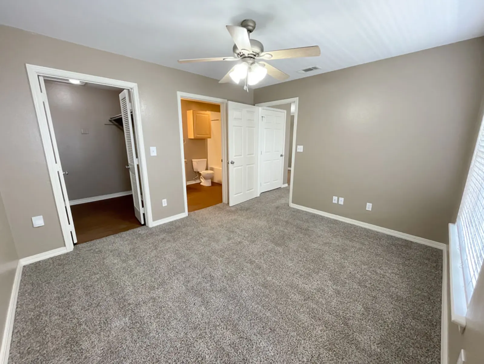 A spacious bedroom featuring beige walls, a ceiling fan, carpeted flooring, and two closets. A door leads to a bathroom, and there are windows allowing natural light to enter.