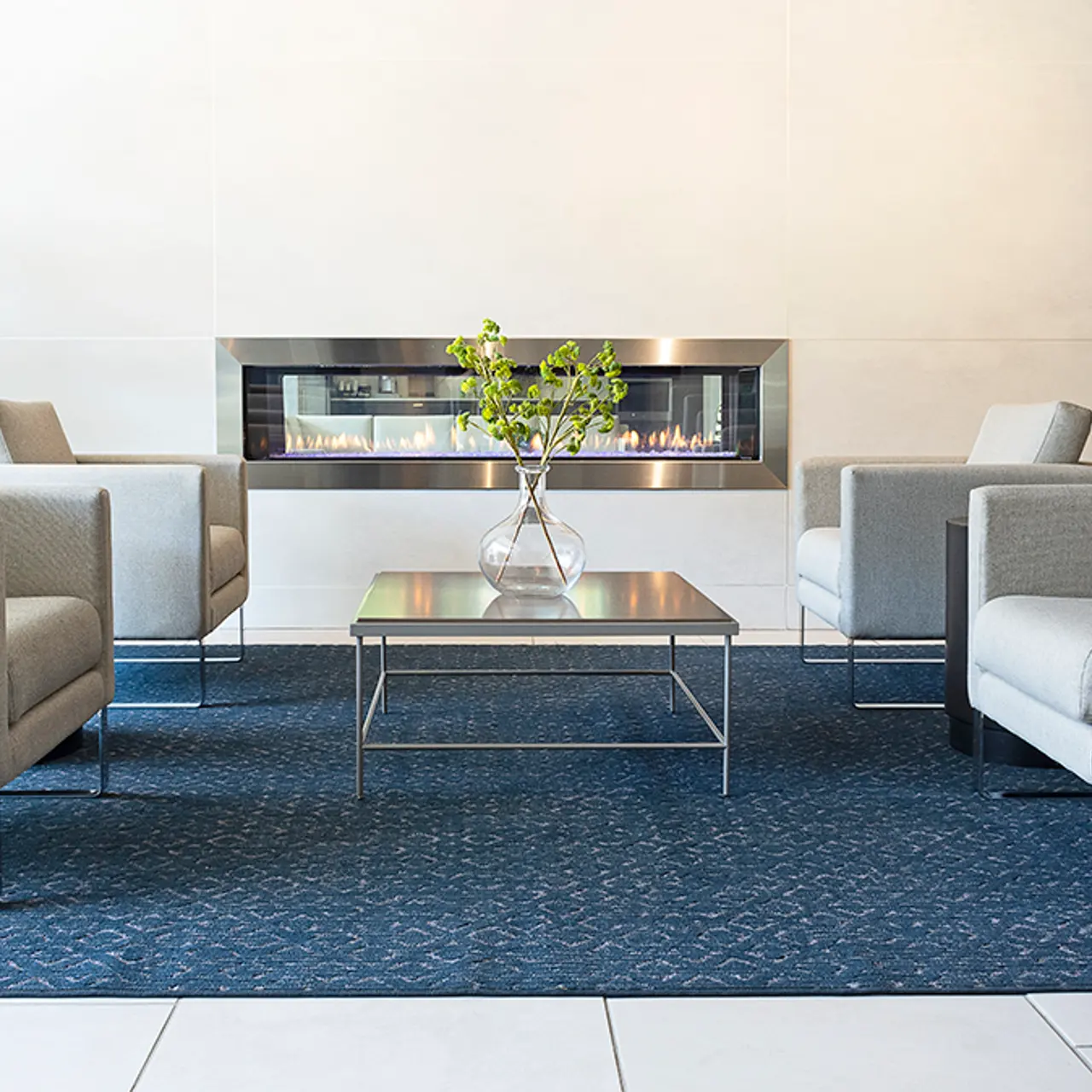 Modern Lounge Area with Fireplace A modern lounge area featuring four light gray armchairs arranged around a glass table, with a decorative vase and greenery on the table. In the background, there's an elegant fireplace mounted on a wall. The floor has light-colored tiles and a blue patterned rug beneath the furniture.