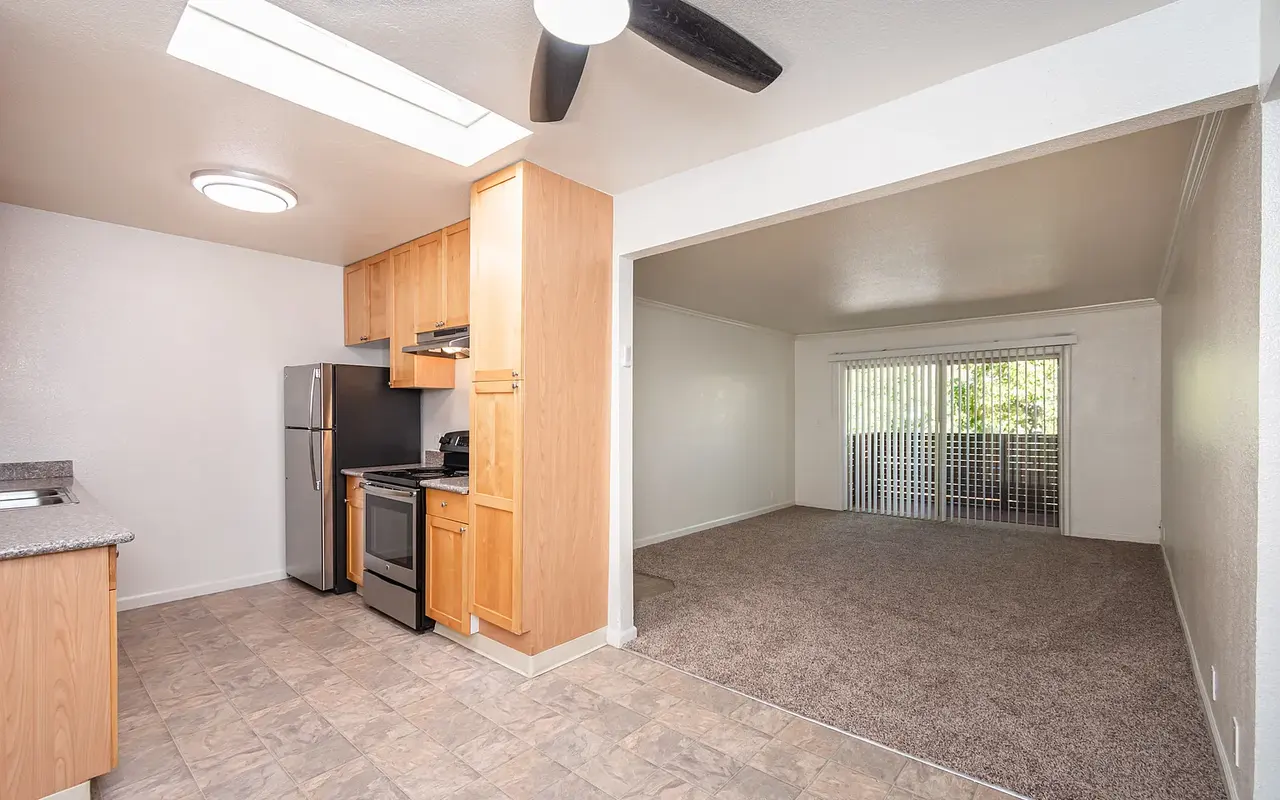 Interior of a cozy apartment featuring a kitchen with wooden cabinets and appliances, leading to a carpeted living area with large windows.