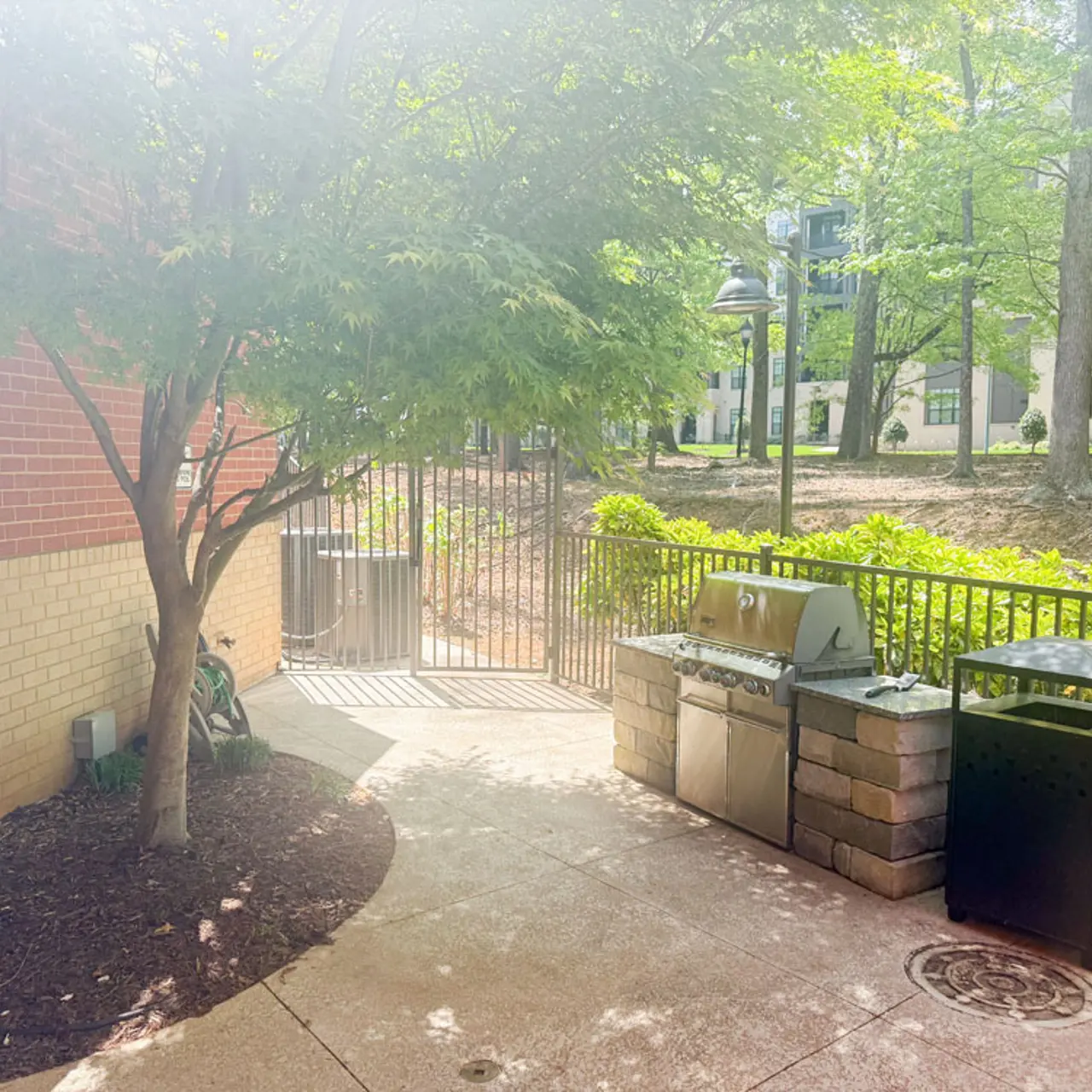 An outdoor grilling area with a tree, grill station, and garbage bin, surrounded by greenery and a gated path.