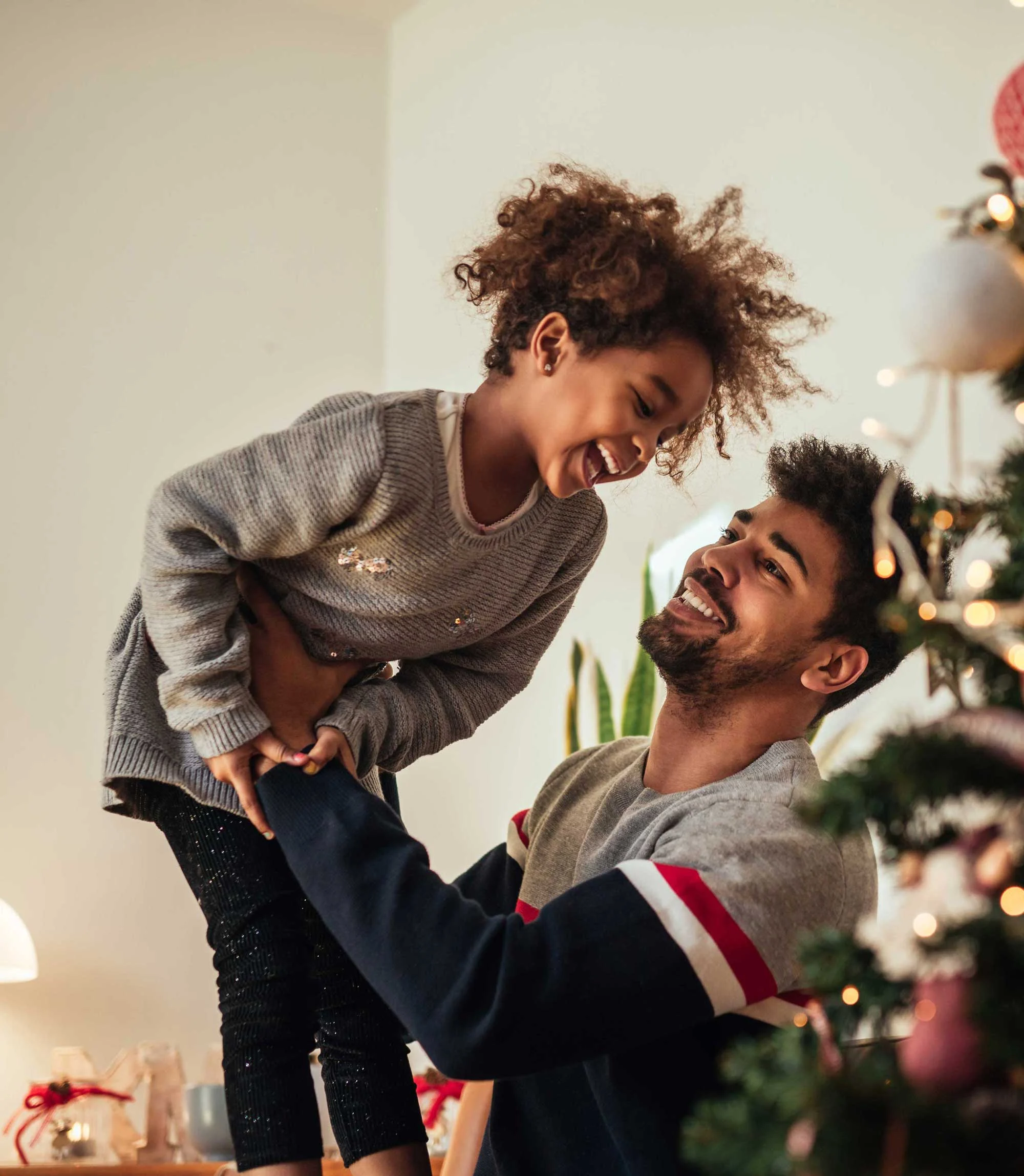 A smiling father holds up his laughing daughter in a festive room decorated for the holidays, with a Christmas tree in the background.