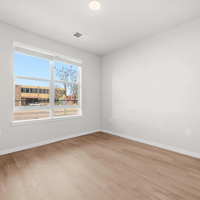 A bright and empty room with light-colored walls and wood flooring. A large window lets in natural light, showing views of an exterior building and trees.