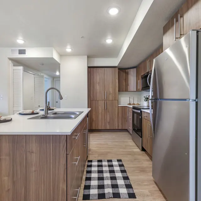 Modern Kitchen Design A modern kitchen featuring wooden cabinets, a stainless steel refrigerator, and a large island with a sink. The floor is a light wood, and there is a black and white checkered rug on the floor. Natural light is accentuating the space.