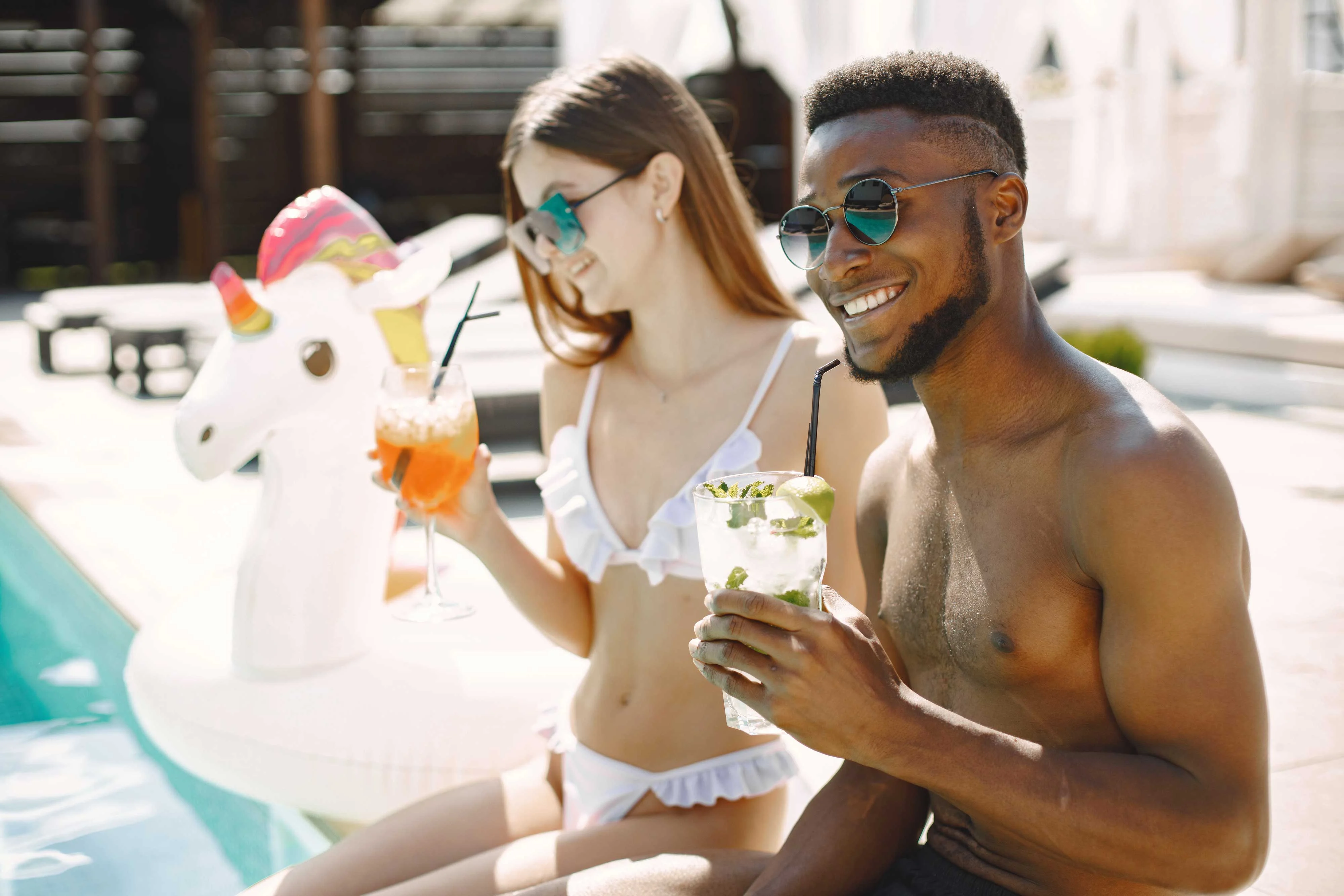 A young couple enjoying drinks by a swimming pool, sitting on the edge with a unicorn float nearby.