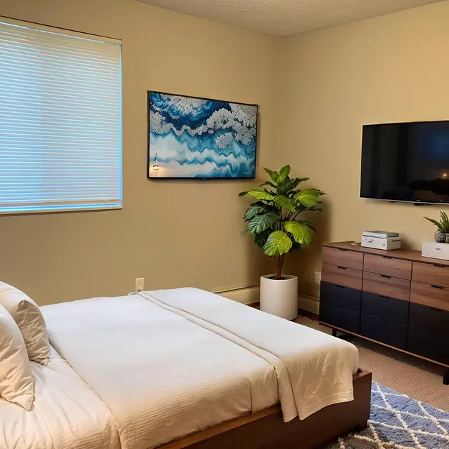 A cozy bedroom featuring a bed with white linens, a TV mounted on the wall, a wooden dresser, and a green plant in the corner. There is a piece of art on the wall depicting blue waves, and a lamp on the bedside table.