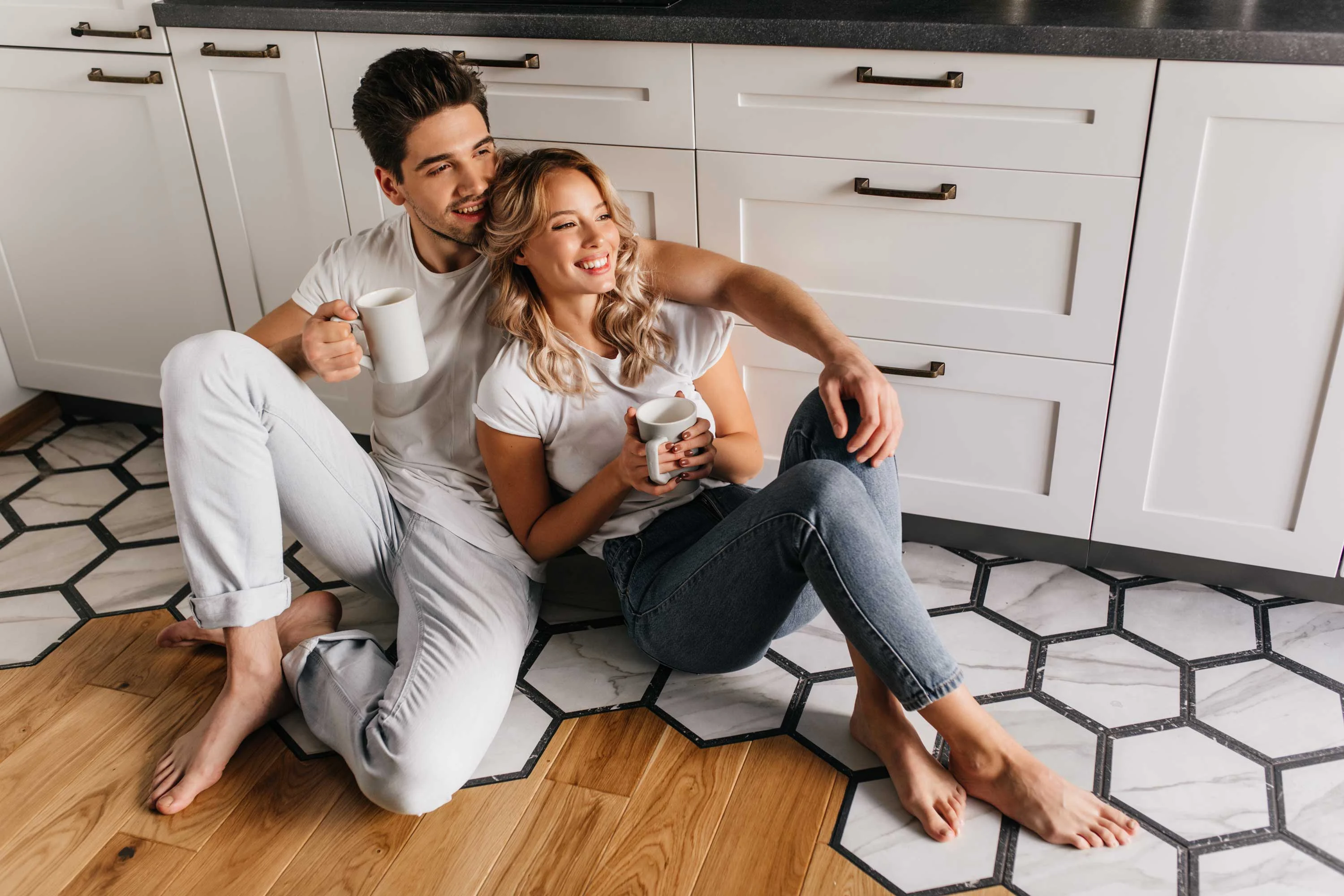 The Gleason A couple sitting on the kitchen floor, holding mugs and smiling at each other. They are dressed in casual clothing, surrounded by a stylish kitchen with distinct tile flooring.