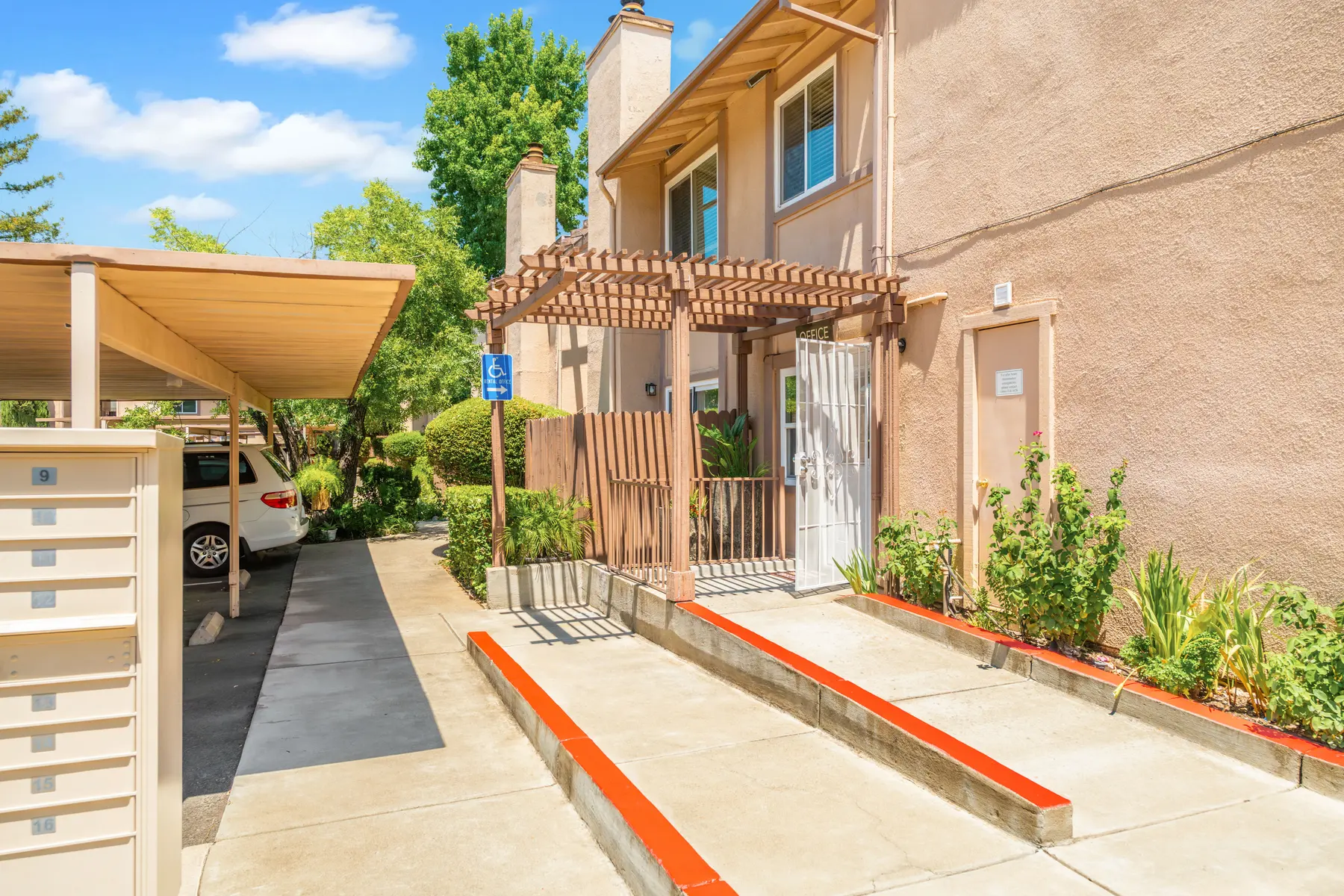 A residential building entrance with a wooden trellis, a ramp, and landscaped greenery.