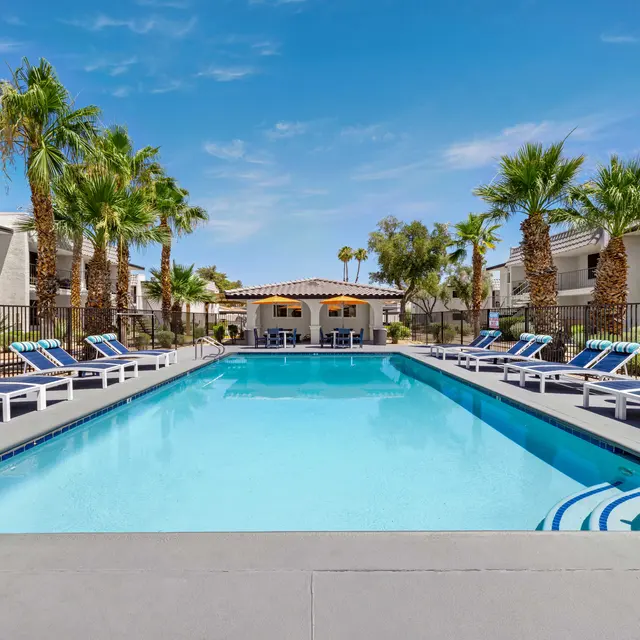 A clear blue swimming pool surrounded by lounge chairs and palm trees in an apartment complex.