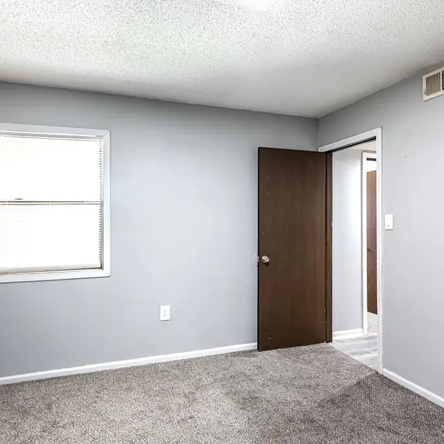 A small, empty bedroom with gray walls and carpet. A window with horizontal blinds is visible on the left, and a closed wooden door is on the right. A door leads to another room at the back.