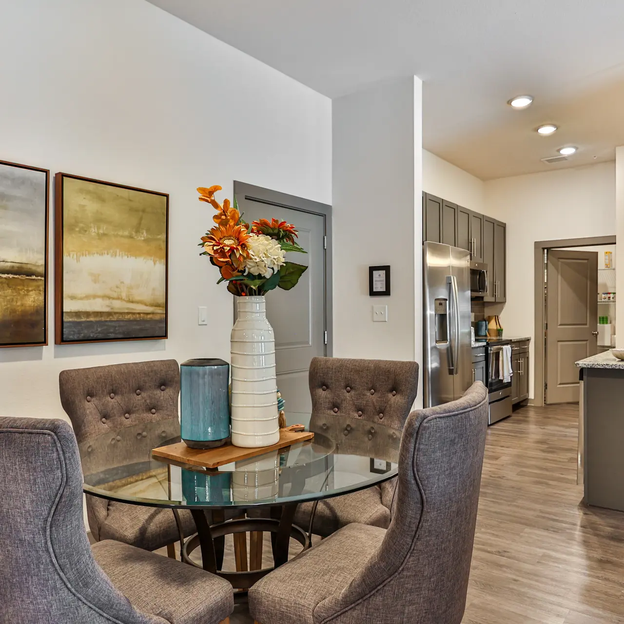 Modern Kitchen and Dining Area A modern dining area featuring a round glass table with four upholstered chairs. A vase of flowers sits at the center of the table, surrounded by two abstract paintings on the wall. The kitchen area is visible in the background, showcasing modern appliances and cabinetry.