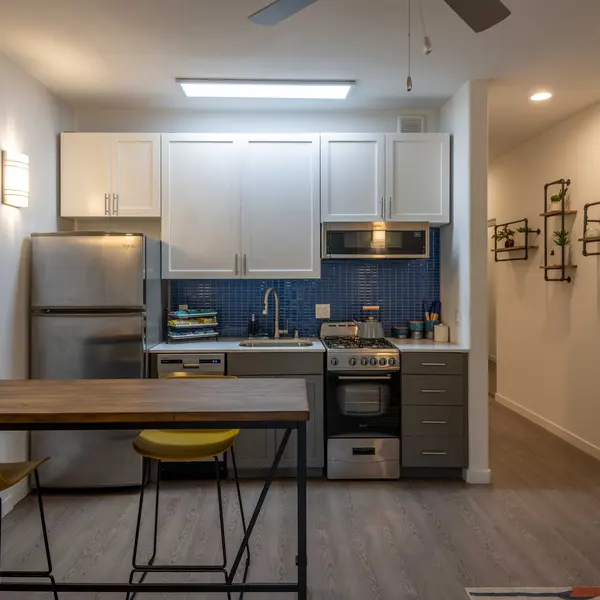 A small modern kitchen featuring a stainless steel fridge, a stove with an oven, and light-colored cabinetry. The kitchen has blue tiled backsplash and a bar table with yellow stools. There are also hanging plants on the wall and a bright light fixture overhead.