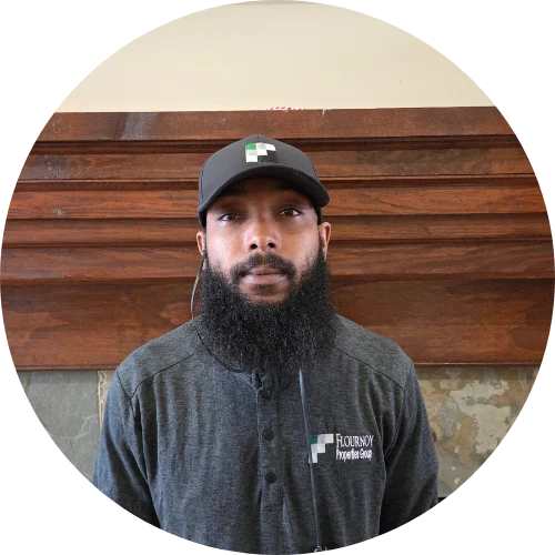 A man with a beard and wearing a black cap and a gray shirt stands in front of a wooden-paneled wall.