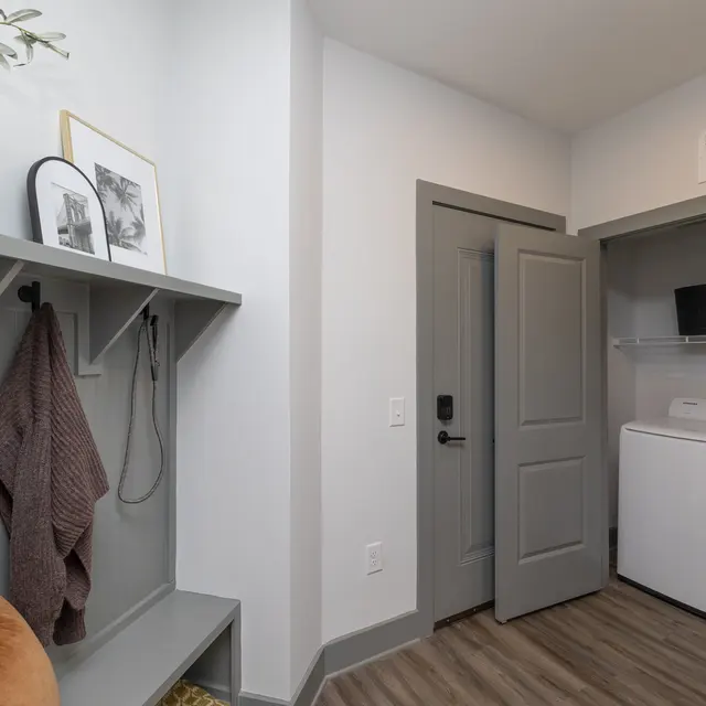 Stylish Laundry Room Interior A modern laundry room featuring a washer and dryer. The room has light gray walls and wooden flooring. There’s a coat rack on the wall with a brown towel hanging. A small potted plant is on a ledge near a decorative picture frame.