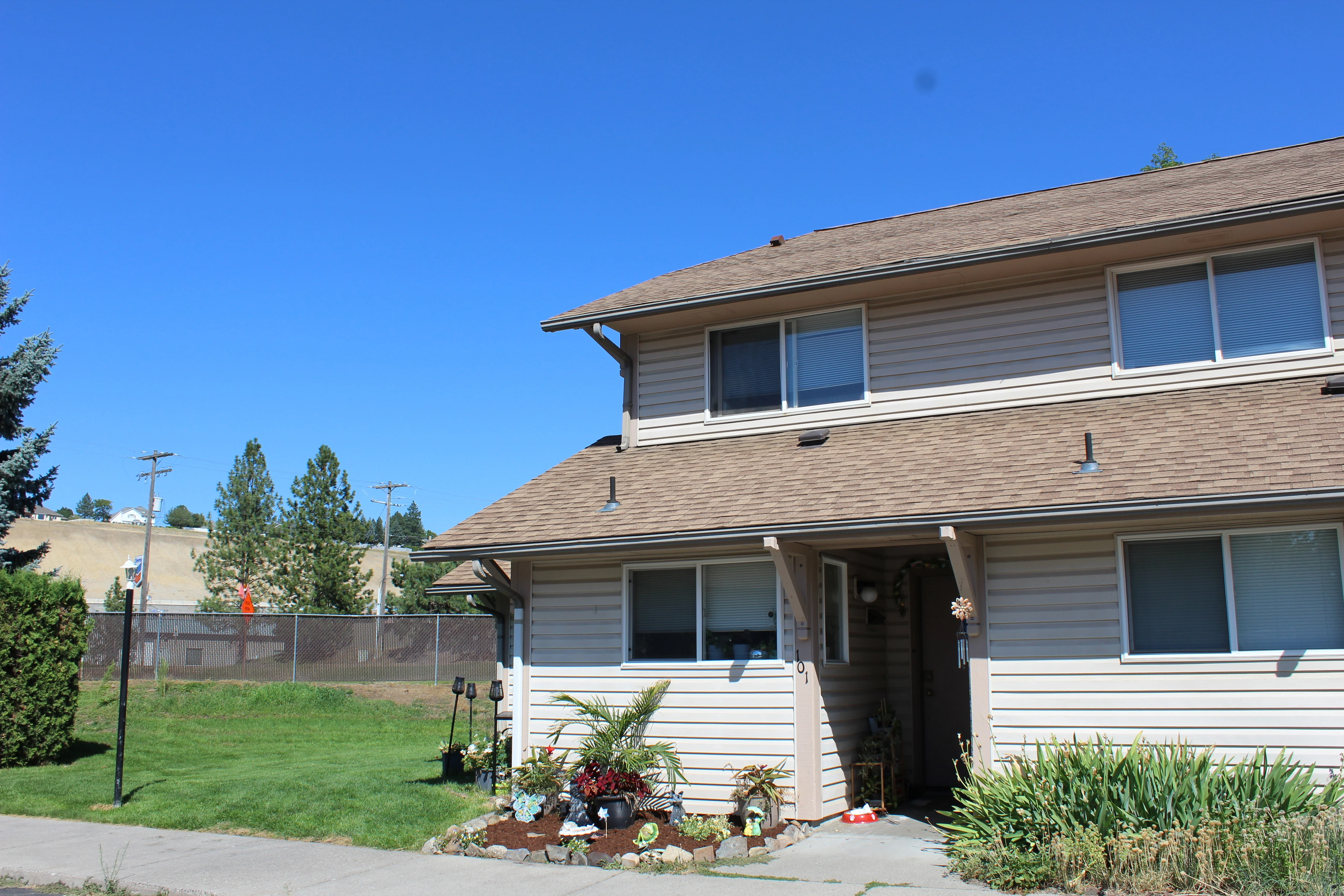 A two-story residential building with a brown exterior and a green lawn in front. It features a small garden with potted plants and has a clear blue sky in the background.
