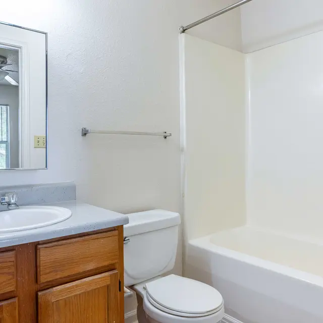A simple bathroom featuring a bathtub, toilet, and a sink with wooden cabinets. A mirror is mounted above the sink and a window is visible in the background.
