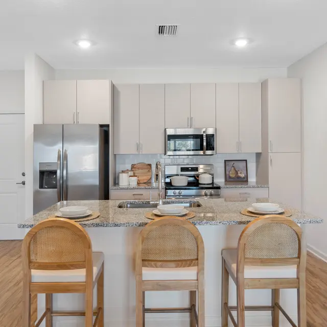 A modern kitchen featuring a central island with three wooden bar stools, stainless steel appliances, and light-colored cabinetry.