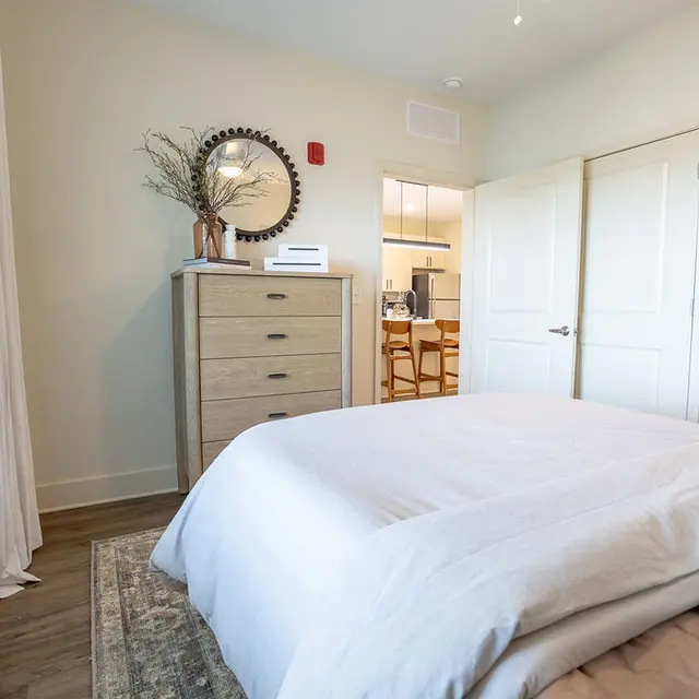 A cozy modern bedroom featuring a beige bedspread, a dresser with decorative items, and a large round mirror above the dresser.