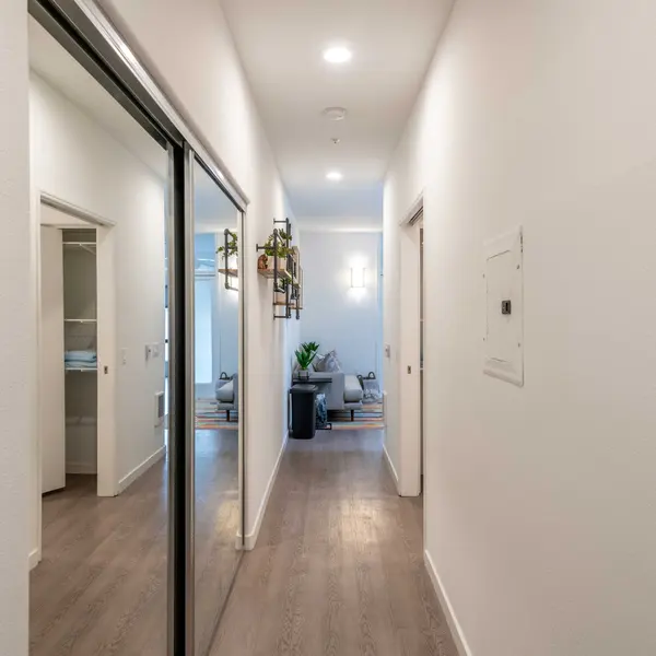 A clean, modern hallway in an apartment with light-colored walls and a wooden floor. There are mirrors on the left side and plants visible through the doorway at the end of the hallway.