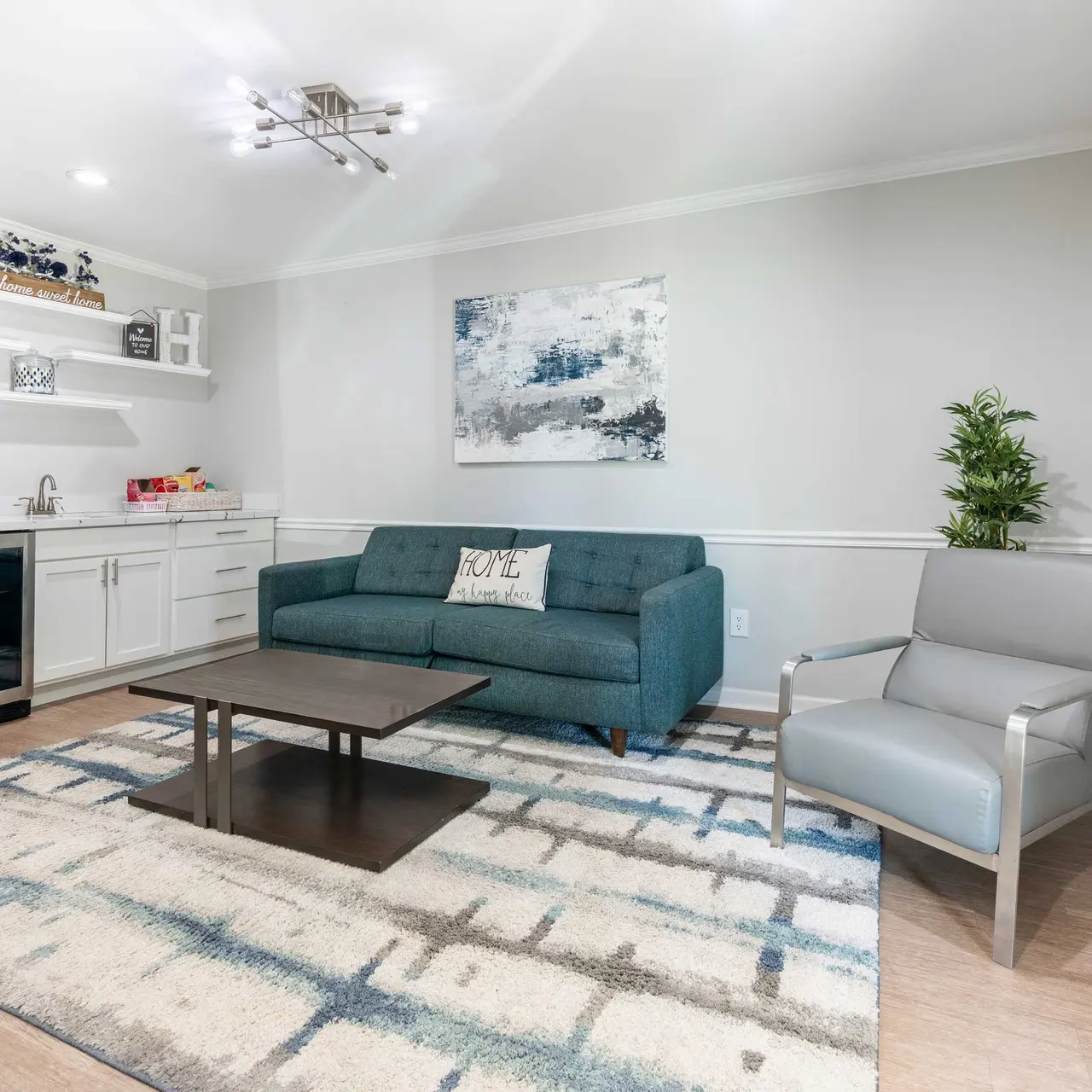 A modern living room featuring a teal couch, gray armchair, and a coffee table on a patterned rug. There is a kitchenette with a coffee maker and a small refrigerator in the background.
