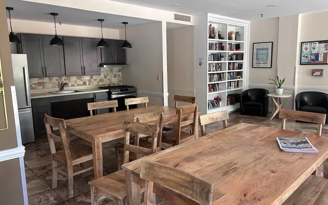 Communal Dining Space A communal dining area featuring wooden tables and chairs, with a kitchen in the background and bookshelves on one side.