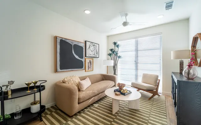 Stylish Modern Living Room A modern living room featuring a beige sectional sofa, a round coffee table, and an accent chair. There are artworks on the wall, a floor lamp beside the sofa, and a potted plant in the corner. The room has a striped rug and illuminated by natural light from large windows with blinds.