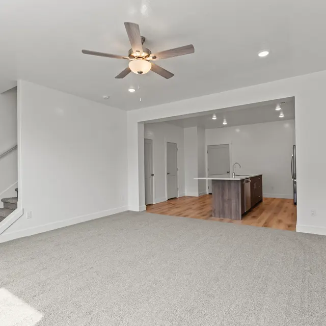 A spacious living room featuring light-colored carpet, a ceiling fan, and a view into a kitchen area with wooden flooring and modern appliances.