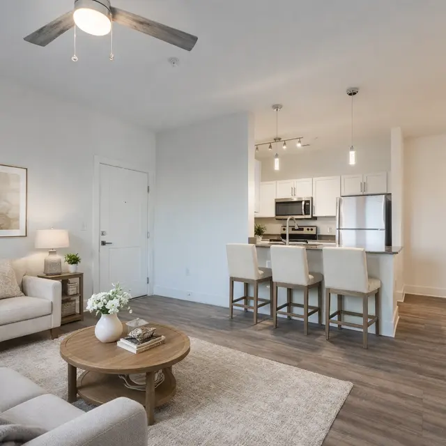 A modern living room area with a beige sofa, a round wooden coffee table, and a decorative plant. In the background, there's an open kitchen with stainless steel appliances and a counter with bar stools.