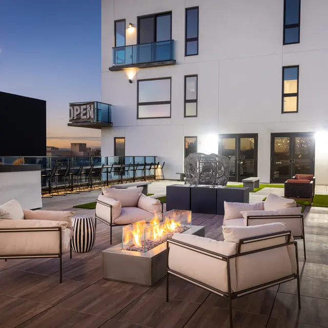 A modern rooftop patio with cozy seating, a fire pit, and dim evening lighting, featuring an open sign in the background.