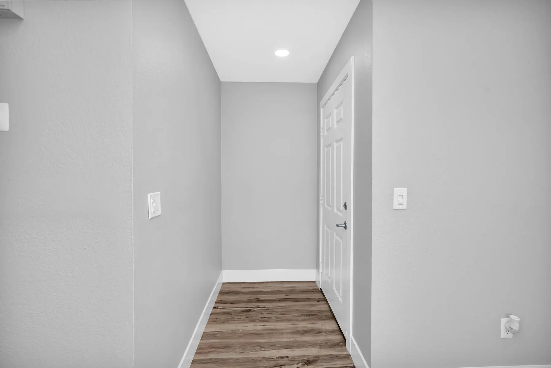 A simple, modern hallway with light gray walls, wood-like flooring, and a closed door on the right side.