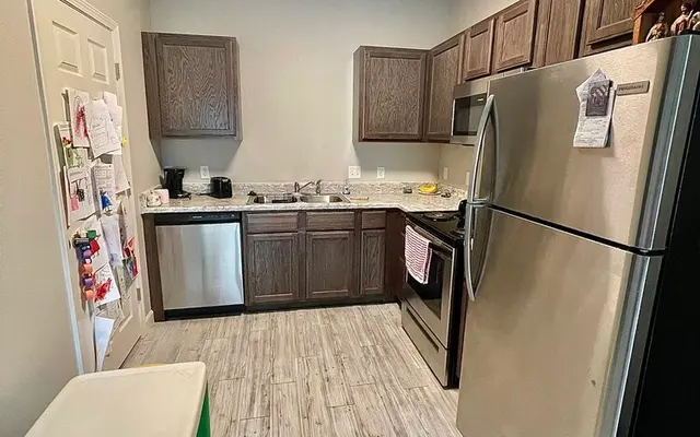 A modern kitchen with light gray walls and dark wood cabinets. The kitchen features stainless steel appliances, including a fridge and a stove. A small white table with green and yellow seats is in the corner. There’s a light source coming from above, giving a bright ambiance.