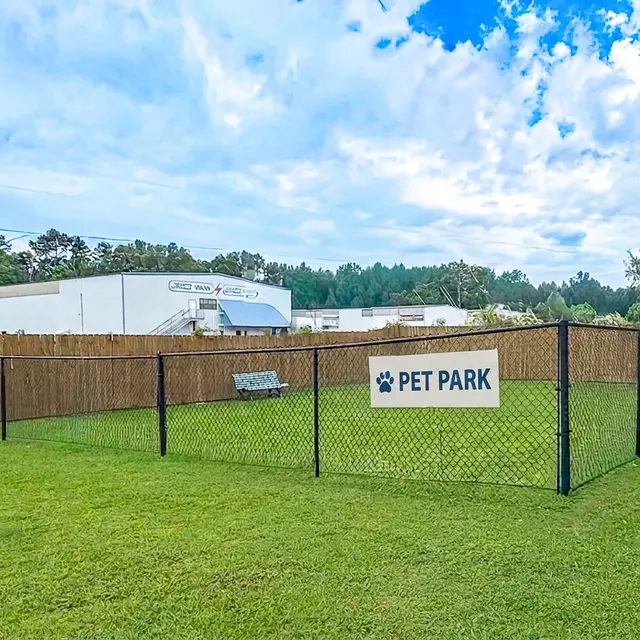 A fenced area labeled 'PET PARK' with a bench inside, surrounded by green grass and a wooden fence. In the background, there are buildings and trees under a partly cloudy sky.