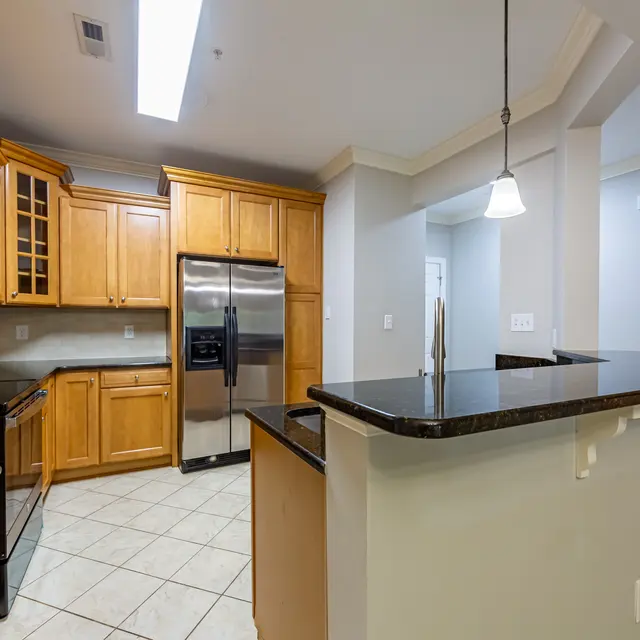 A modern kitchen featuring wooden cabinets, stainless steel appliances, and granite countertops. The kitchen has a bar area with stools and a light fixture above. Tile flooring is visible throughout the space.