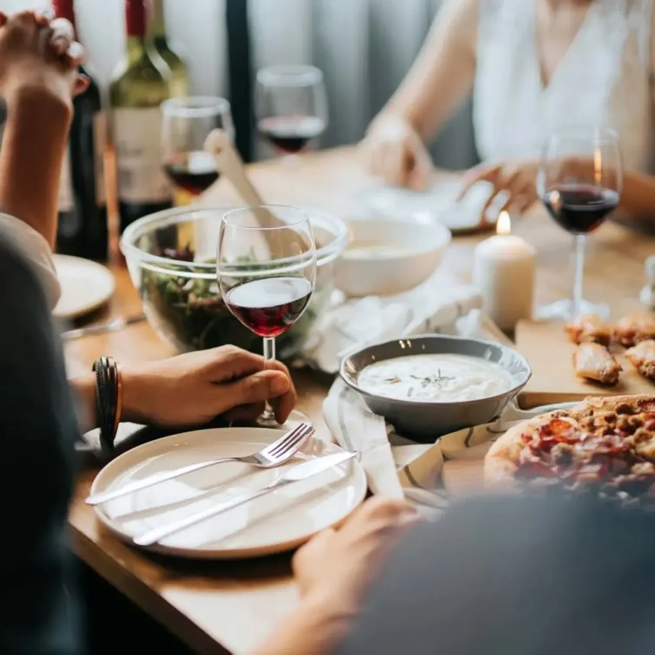 A group of people enjoying a dinner gathering with a variety of dishes on the table, including pizza, salad, and appetizers, alongside glasses of red wine.