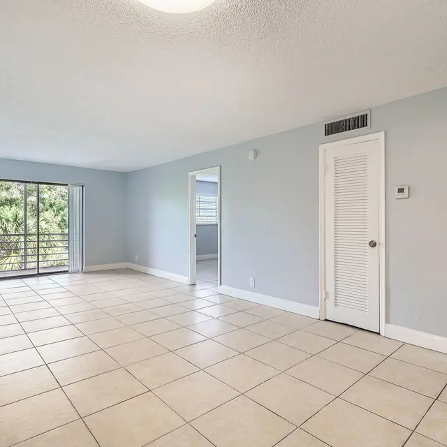 A spacious and empty living room with light blue walls and tiled flooring, featuring a large window with sliding glass doors that opens to a balcony area. A white door is visible on the right side leading to another room.