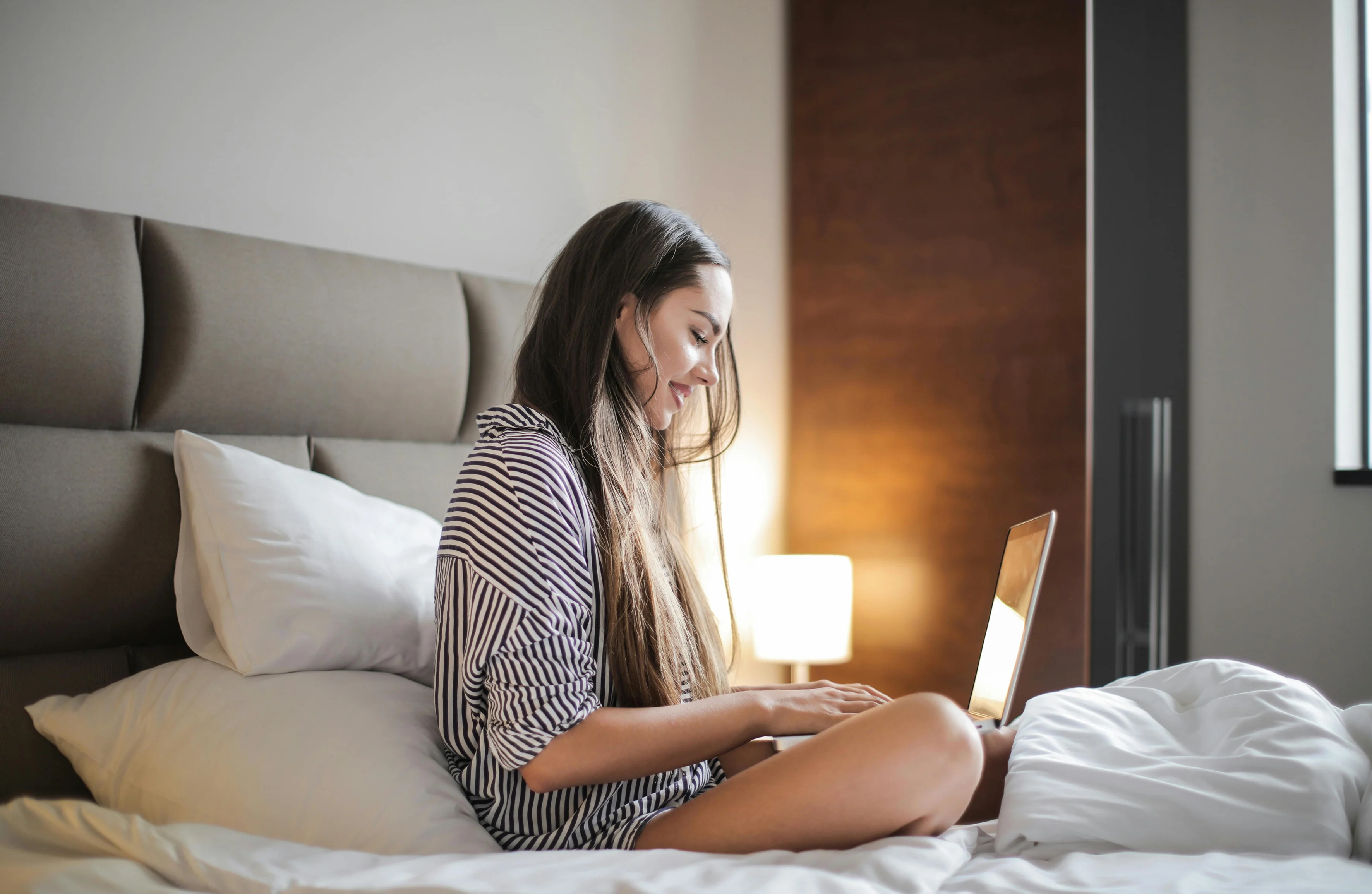 Working from Bed: A Cozy Morning Routine A young woman sitting on a bed, focused on a laptop. The room has a cozy atmosphere with natural light and simple decor.