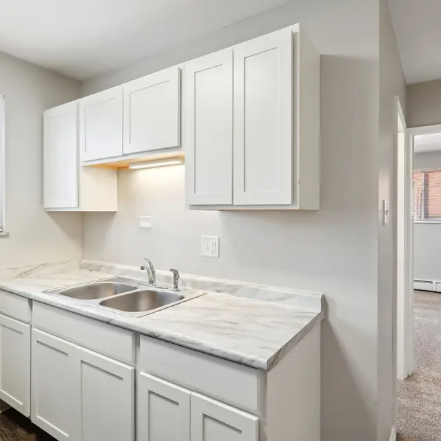 A modern kitchen featuring white cabinets, a marble countertop, and a double sink. Natural light comes in through a window, illuminating the space. A doorway leads to another room in the background.