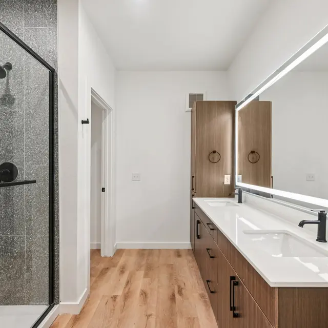 A modern bathroom featuring a glass shower enclosure, a double vanity with sink and sleek fixtures, and wooden accents on the cabinetry. The space has bright lighting and elegant flooring.