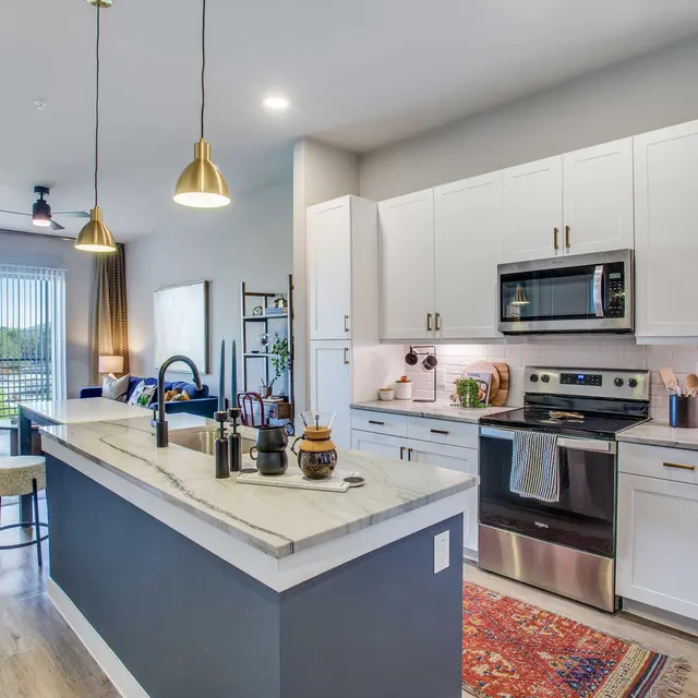 A modern kitchen with a large island, stainless steel appliances, white cabinets, and pendant lighting. There is a cozy living space visible in the background with windows overlooking a cityscape.