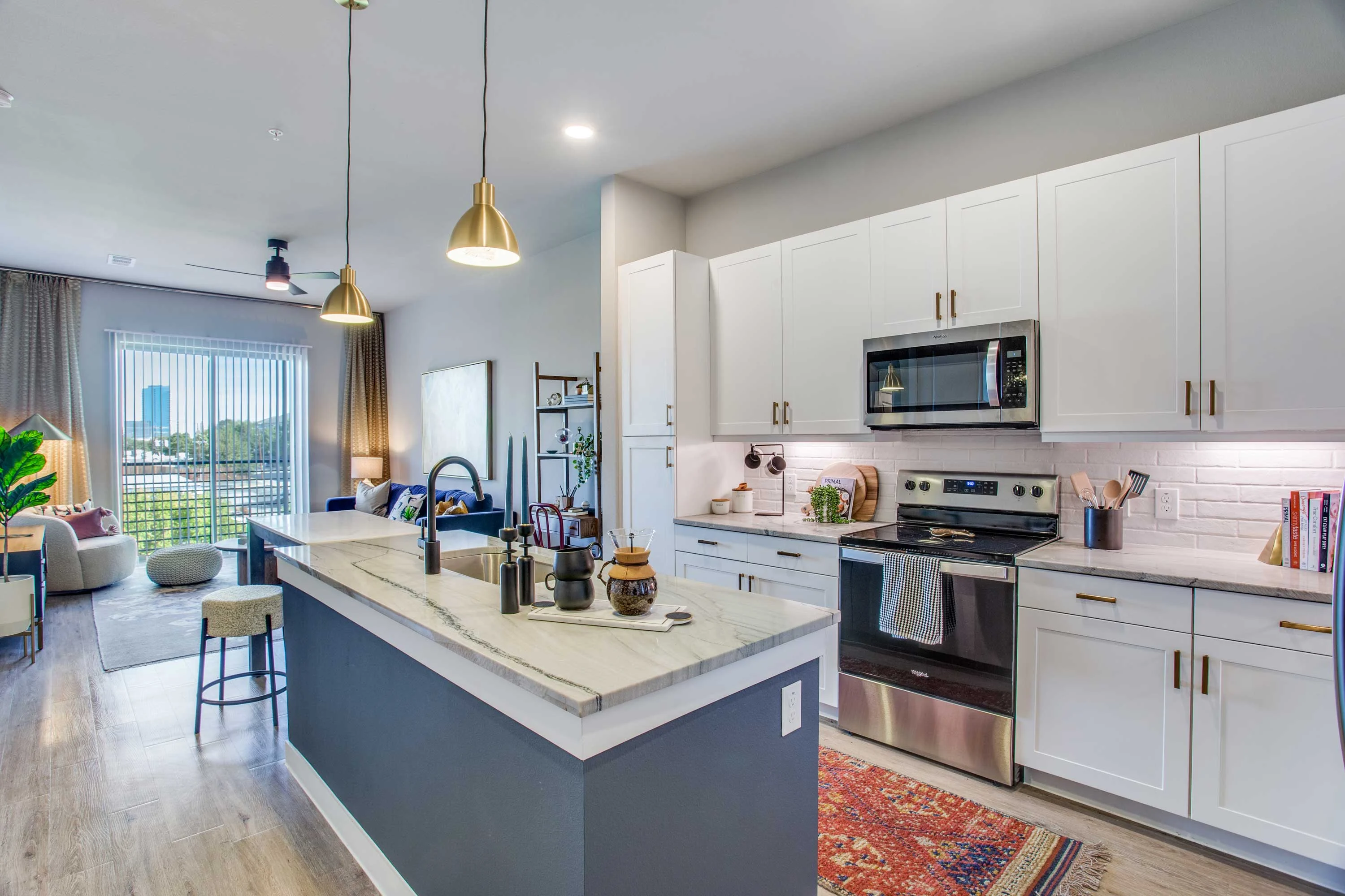 A modern kitchen with a large island, stainless steel appliances, white cabinets, and pendant lighting. There is a cozy living space visible in the background with windows overlooking a cityscape.