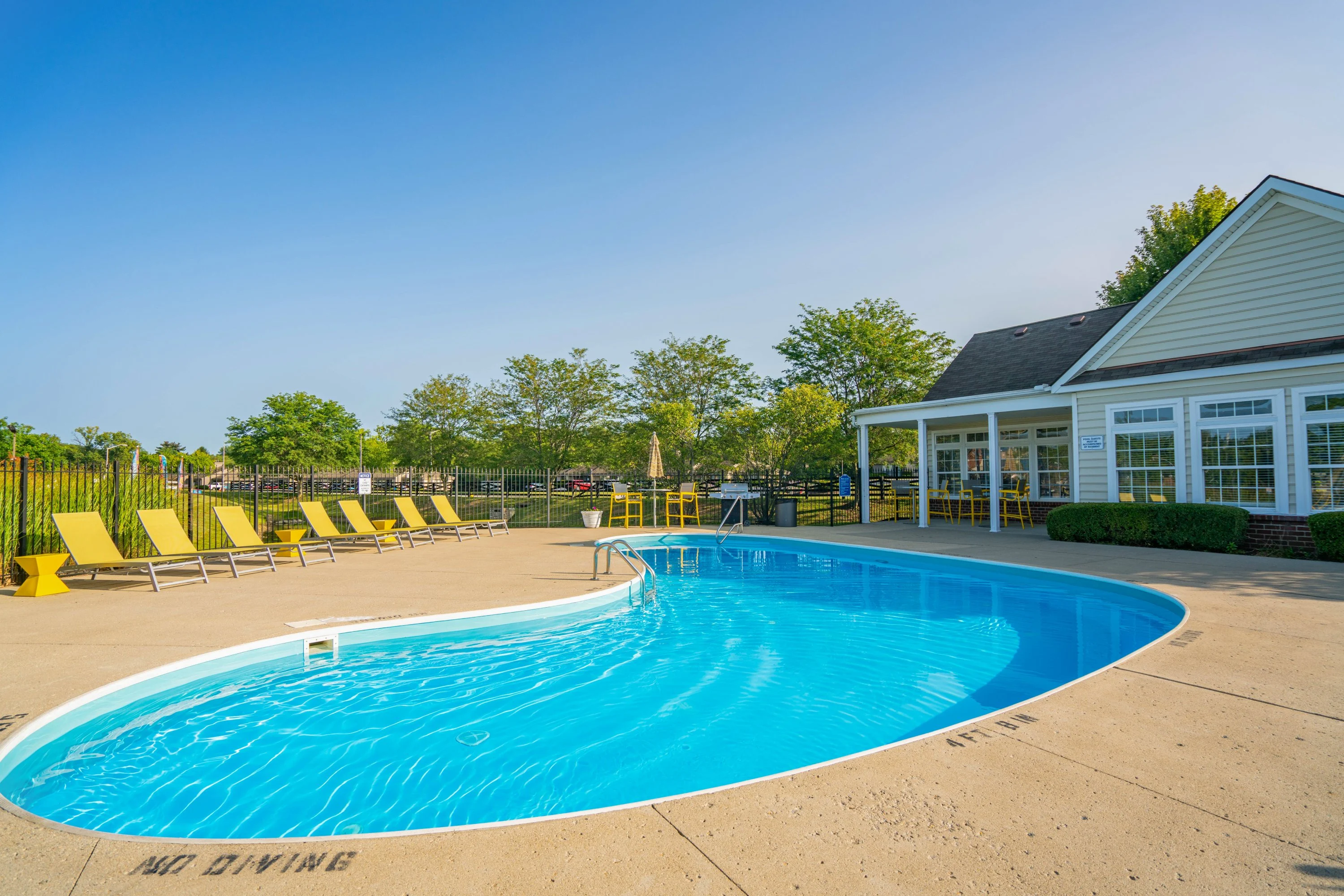 Community Pool Area A swimming pool with a clear blue water, surrounded by yellow lounge chairs and a community building in the background.