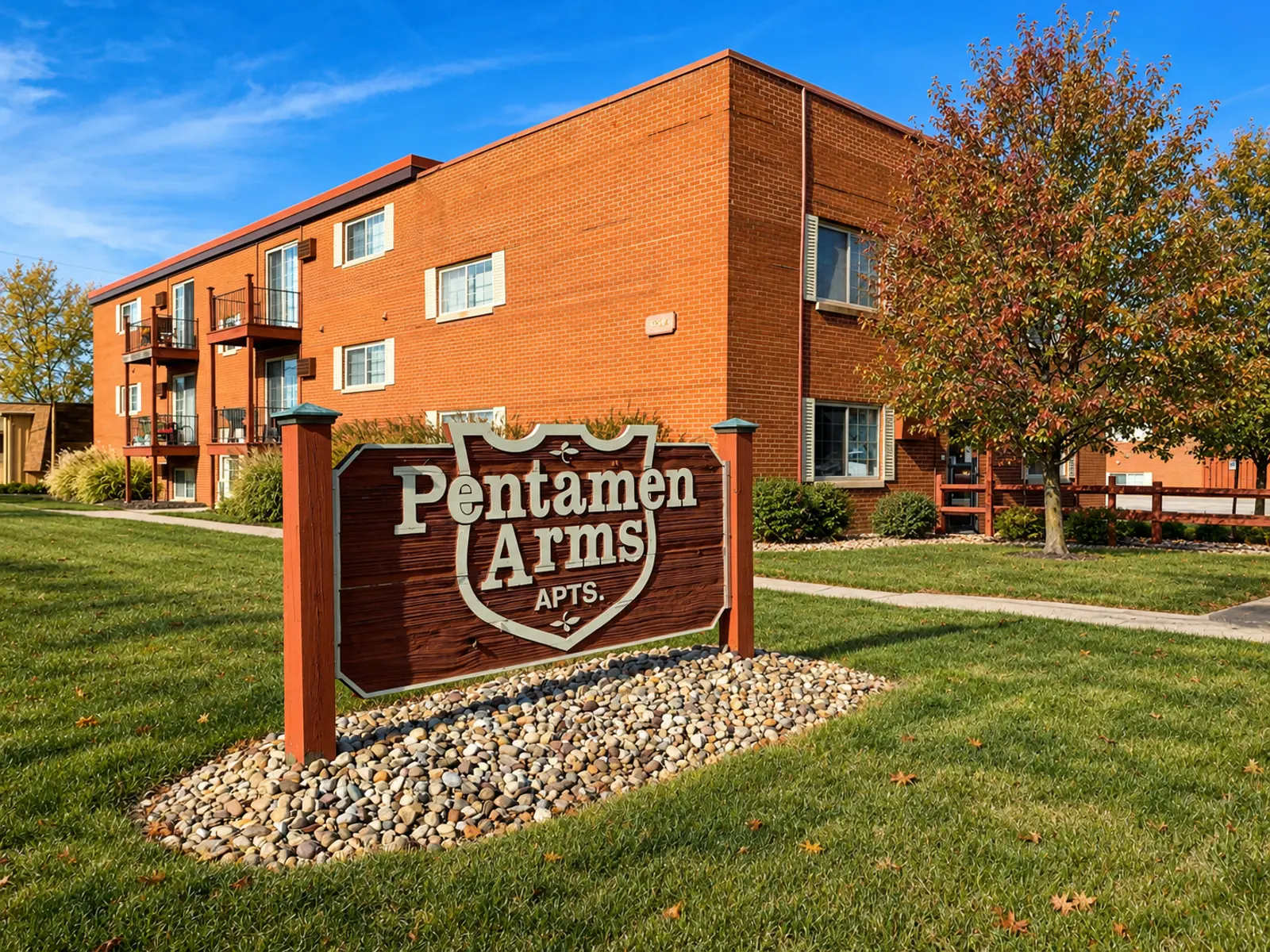 A red-brick apartment building with a sign that reads 'Pentamen Arms Apts.' in a grassy area.
