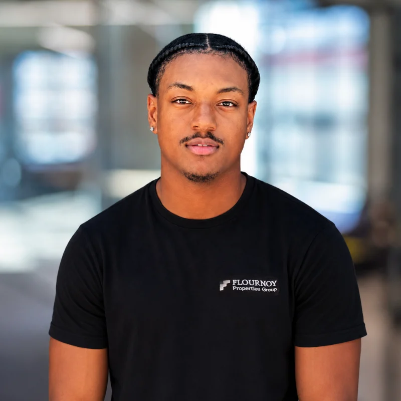 A professional portrait of a man wearing a black t-shirt with a company logo, standing outdoors with soft focus in the background.