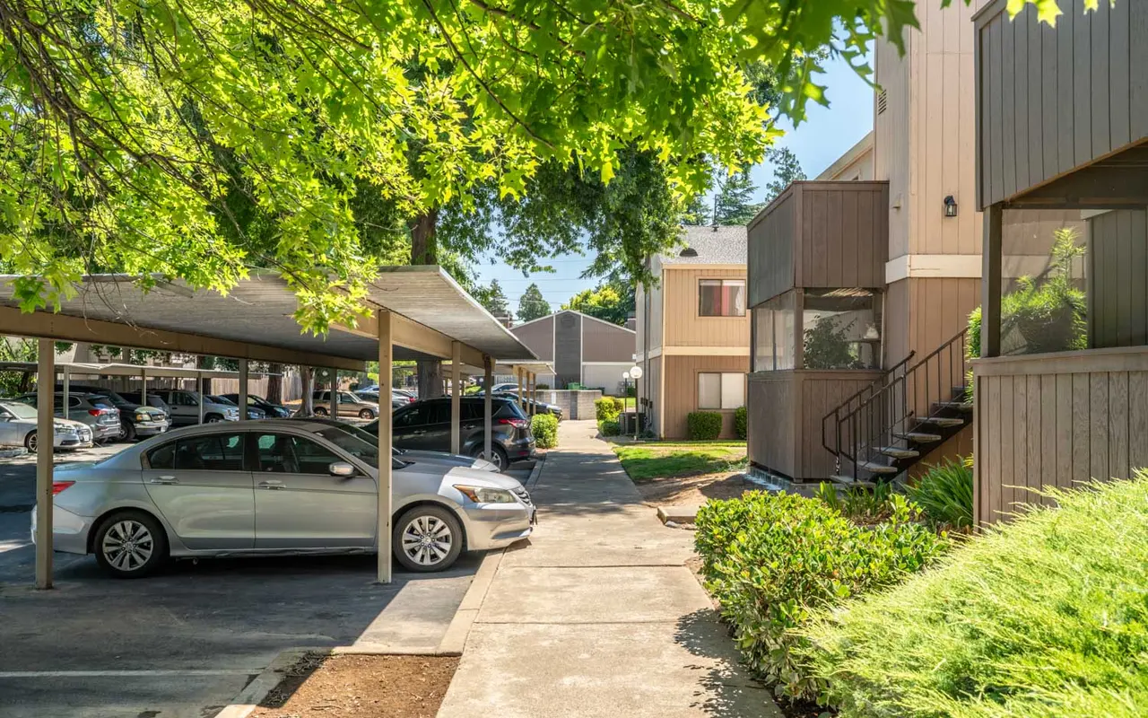 A view of a parking area in an apartment complex, featuring covered parking spaces and a well-maintained sidewalk leading to a building entrance.