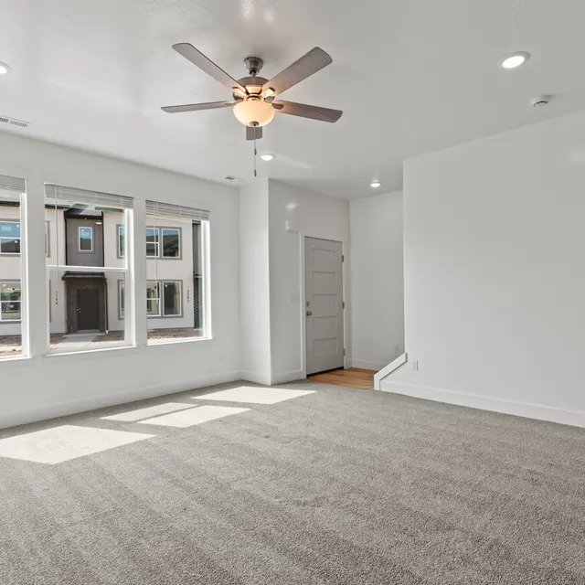 A spacious open living room with large windows, a ceiling fan, and light-colored carpet flooring.